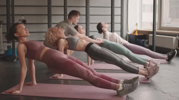 Colpo completo di quattro giovani donne di razza mista e caucasica che fanno tavola inversa su tappetini da yoga in palestra di giorno, l'allenatore maschio li aiuta con la tecnica - Filmati, video