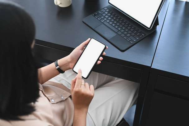 Накладний знімок молодої жінки сидить і її офісний стіл і тримає мобільний телефон з білим екраном
. - Фото, зображення