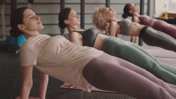 Mediano largo de cuatro mujeres jóvenes deportes magras levantándose en tablón inverso en esteras de yoga en el gimnasio durante el día - Imágenes, Vídeo