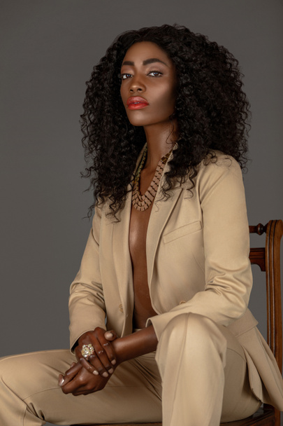 Porträt einer sinnlichen jungen schwarzen Frau mit langen lockigen schwarzen Haaren und schönem Make-up, die allein auf einem Holzstuhl in einem Studio mit grauem Hintergrund sitzt und einen beigen Anzug und Schmuck trägt.. - Foto, Bild