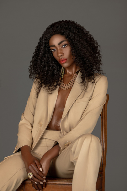 Retrato de una joven negra fuerte con el pelo largo y rizado negro y hermoso maquillaje sentada sola en una silla de madera en un estudio con un fondo gris con un traje beige y joyas. - Foto, imagen