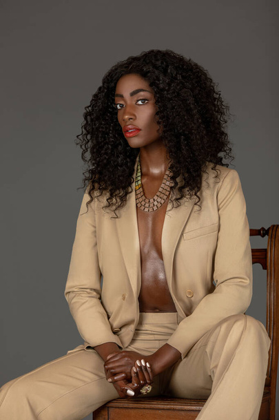 Porträt einer attraktiven jungen schwarzen Frau mit langen lockigen schwarzen Haaren und schönem Make-up, die allein auf einem Holzstuhl in einem Studio mit grauem Hintergrund sitzt und einen beigen Anzug und Schmuck trägt.. - Foto, Bild