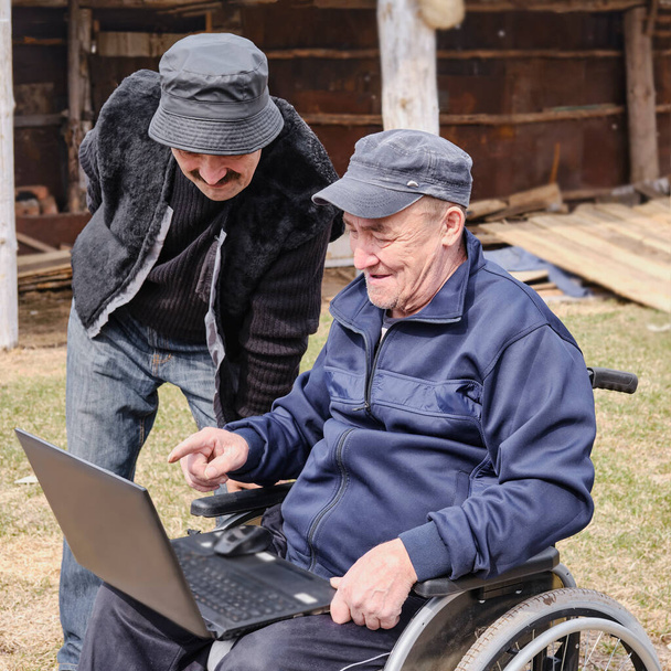 Glücklicher älterer Mann im Rollstuhl im Hof seines Hauses, sein Freund lächelt und blickt auf einen Laptop. - Foto, Bild