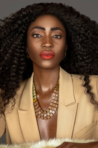 Nahaufnahme Porträt einer starken schwarzen Frau mit langen lockigen schwarzen Haaren und schönem Make-up, die allein auf einem Pelz in einem Studio mit grauem Hintergrund posiert und einen beigen Anzug und Schmuck trägt. - Foto, Bild