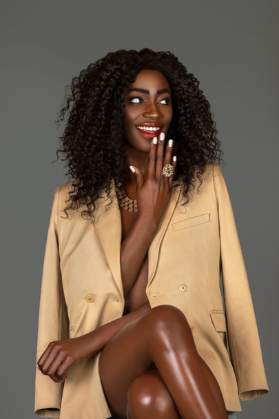 3/4 portrait of a happy black woman with long curly black hair and beautiful makeup sitting by herself in a studio with gray background just wearing a beige jacket and jewelry. - Photo, Image
