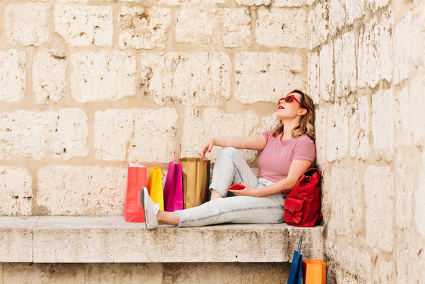 Mujer con gafas de sol rojas descansando en la calle de su ciudad rodeada de coloridas bolsas concepto adicto a las compras, ventas y ofertas. Estilo de vida. - Foto, Imagen