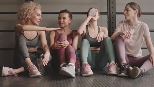 Foto completa de cuatro deportistas magras de raza mixta y caucásicas apoyadas en la pared sentadas en el suelo en el gimnasio, bebiendo agua de las botellas, hablando y sonriendo después de un duro entrenamiento - Metraje, vídeo