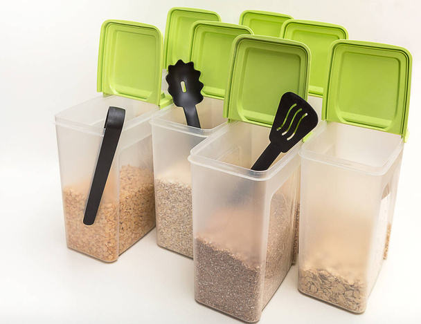 Various cereals - wheat, peas, buckwheat, millet, oatmeal, barley  in open plastic containers with green lids for bulk products and kitchen accessories on a white background, horizontal, side view, diagonal - Photo, Image