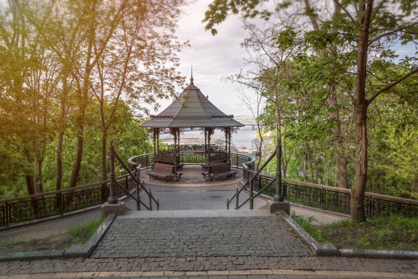 όμορφο πρωινό πάρκο και ένα σφυρήλατο σιδερένιο κιόσκι με ξύλινα παγκάκια για χαλάρωση. Ένα γραφικό κατάστρωμα παρατήρησης με θέα την πόλη του Κιέβου και του ποταμού Δνείπερου. Πάρκο Volodymyrska Girka στο Κίεβο - Φωτογραφία, εικόνα