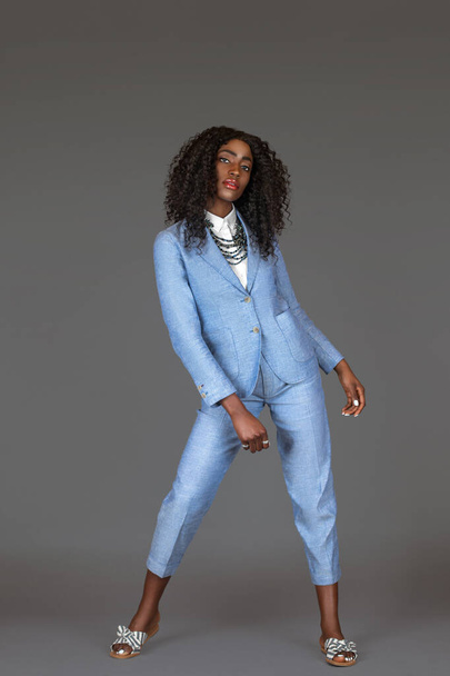 Portrait of a tall sensual black woman with long curly black hair and beautiful makeup posing by herself in a studio with gray background wearing a blue suit with striped sandals and jewelry. - Photo, Image