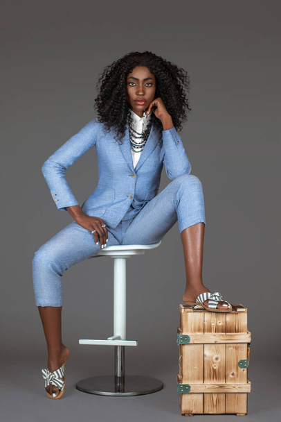 Portrait d'une femme noire sexy avec de longs cheveux noirs bouclés et un beau maquillage assis toute seule sur une chaise dans un studio avec un fond gris portant un costume bleu avec des sandales à rayures et des bijoux. - Photo, image