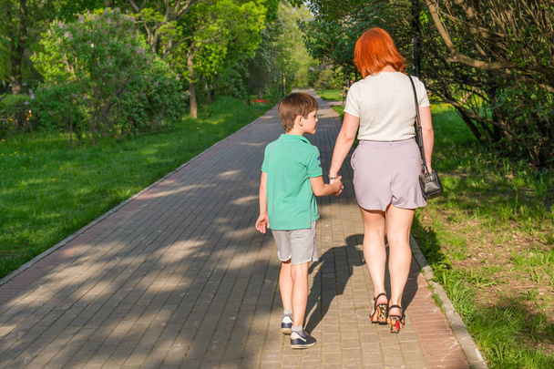Young woman with red hair in shorts and her son are walking along path of city park. Photographed from the back. Mom and son are holding hands. Summer walks with children. - Foto, imagen