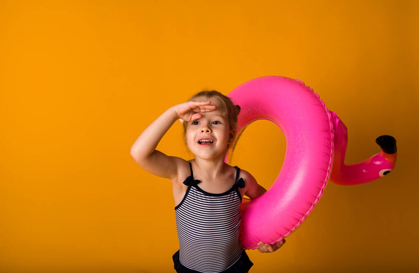 портрет маленькой блондинки в полосатом купальнике, держащей розовый надувной круг с фламинго и глядя на желтый фон с пространством для текста. Летние каникулы - Фото, изображение