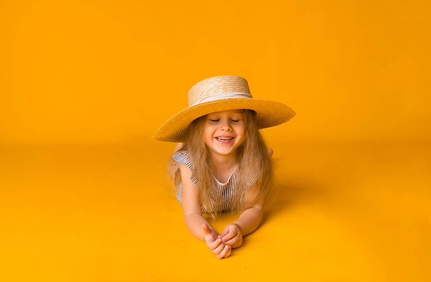 lachend blond meisje met een strohoed ligt op een gele achtergrond met ruimte voor tekst. Zomer achtergrond - Foto, afbeelding