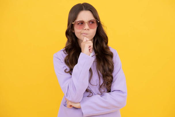 thoughtful kid with curly hair in sunglasses. beauty and fashion. female fashion model. pretty look of young thinking girl in jacket. portrait of stylish child on yellow background. - Photo, Image