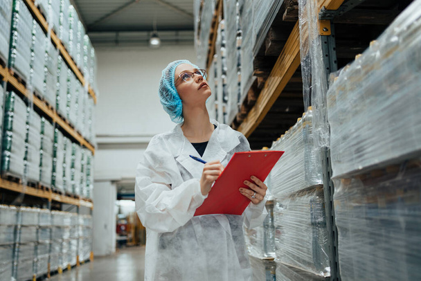 Female worker in protective workwear working in medical supplies research and production factory and checking packed canisters of distilled water before shipment. Inspection quality control. Industrial warehouse. - Foto, immagini