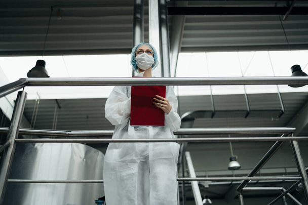 Female worker in protective workwear working in medical supplies research and production factory. She is standing on control bridge and watching at production line. Inspection quality control. - 写真・画像