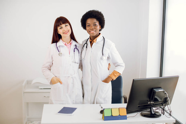 Pretty two young multiethnical women doctors, African and Caucasian, wearing lab coats and stethoscopes, posing embracing each other, standing behind the desk with computer in modern clinic. - 写真・画像