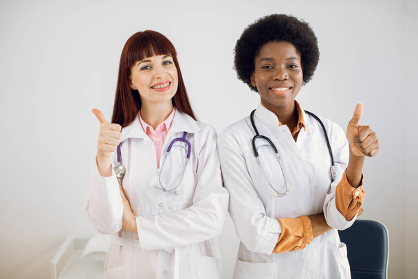 Medizinische Hilfe, Versicherung, Gesundheitsfürsorge und Medizinkonzept. Zwei multiethnische Ärztinnen stehen als Team mit erhobenen Daumen in einem Krankenhausbüro und lächeln in die Kamera. - Foto, Bild