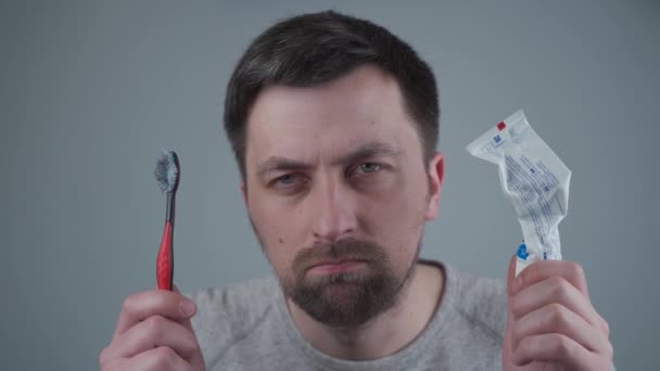 El hombre está molesto por no comprar productos de higiene bucal a tiempo y tenerlos en sus manos pasta de dientes vacía y utiliza cepillo de dientes en el estudio sobre fondo gris. Medios para limpiar los dientes y la boca - Imágenes, Vídeo
