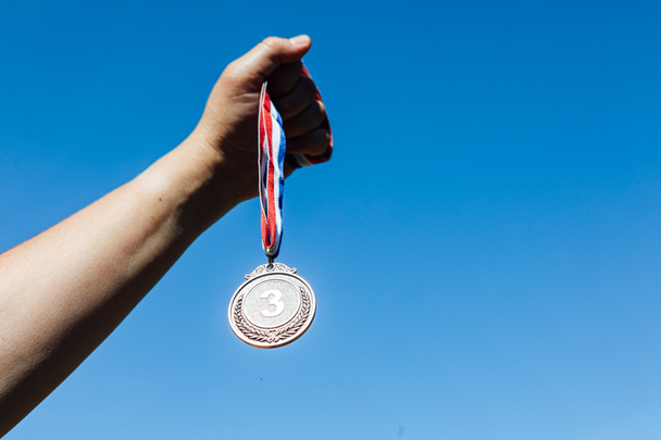 A hand holds up a third-place bronze medal, with the sky in the background. Victory concept - Photo, Image