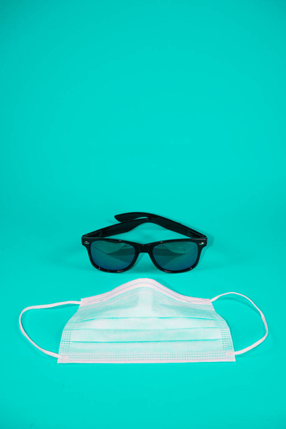 Retro Zwarte plastic zonnebril op turquoise ondergrond. Met een chirurgisch masker voor. Verwoeste zomer door het idee van een corona virus uitbraak. Minimaal abstract zomerconcept. met kopieerruimte. - Foto, afbeelding