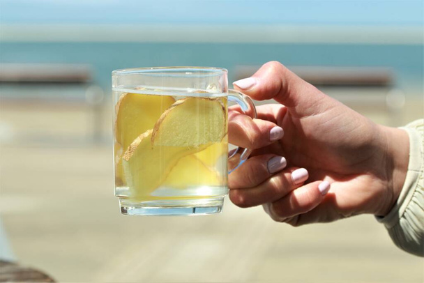 Τσάι τζίντζερ με φέτες τζίντζερ που κρατιούνται από ένα χέρι μπροστά από μια παραλία - Φωτογραφία, εικόνα