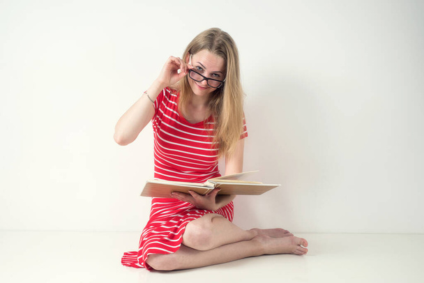 Lány szőke egy piros csíkos ruha szemüveggel és egy könyvet a fehér háttér. - Fotó, kép