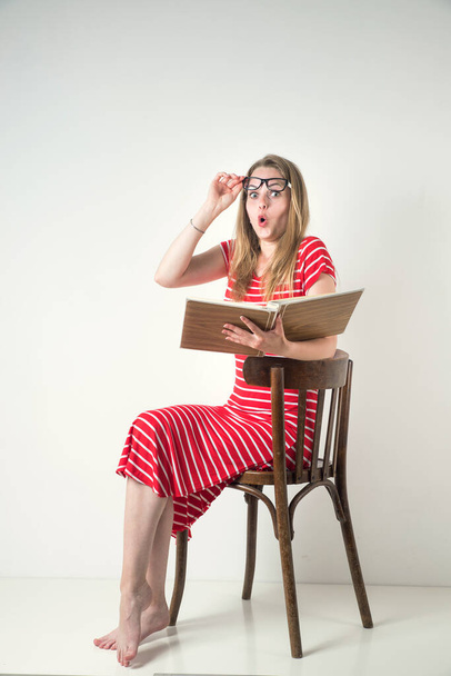 Menina loira em um vestido listrado vermelho com óculos e um livro sobre um fundo branco. - Foto, Imagem