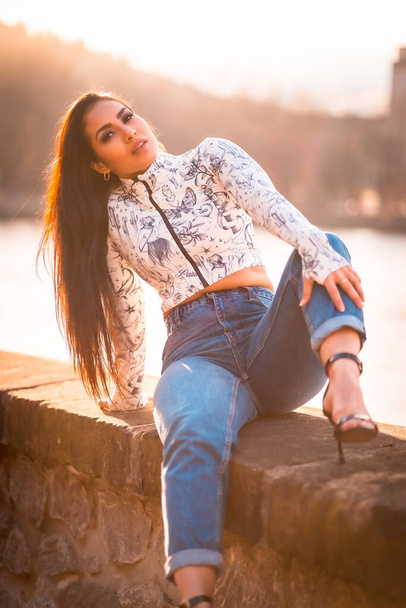 A brunette Latin girl in the city at sunset by the river with the sun in the background. Spring lifestyle - Fotografie, Obrázek
