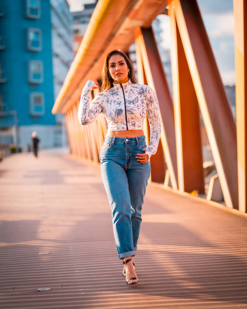 A brunette Latin girl in a jeans in the city at sunset in an orange jumper. Walking across the bridge - 写真・画像