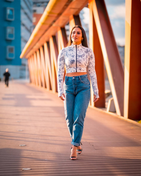 A brunette Latin girl in a jeans in the city at sunset in an orange jumper. Walking across the bridge - Photo, Image