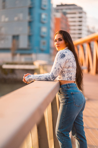 A brunette Latin girl in jeans in the city at sunset. On a bridge looking at the city river - Foto, immagini
