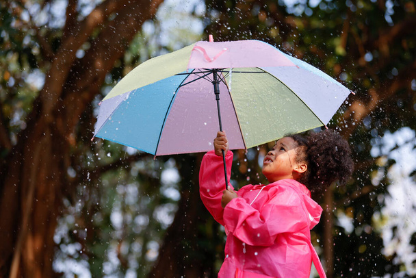 Щаслива смішна дитина кучеряве чорне волосся з парасолькою під осіннім душем. Дівчина носить рожевий плащ і насолоджується дощем. Дитина грає на природі на відкритому повітрі
. - Фото, зображення