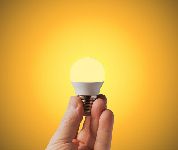 Ilık sarı arka planda bir adamın ellerinde parlayan LED lamba. Enerji tasarrufu kavramı. Boşluğu kopyala. - Fotoğraf, Görsel