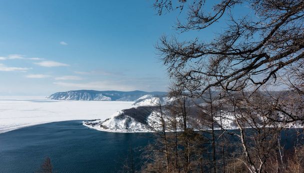 Widok z góry na granicę między białym lodem zamarzniętego jeziora Baikal a błękitną wodą źródła niezamarzniętej rzeki Angara. Góry przeciwko niebu. Gołe gałęzie drzew na pierwszym planie. - Zdjęcie, obraz