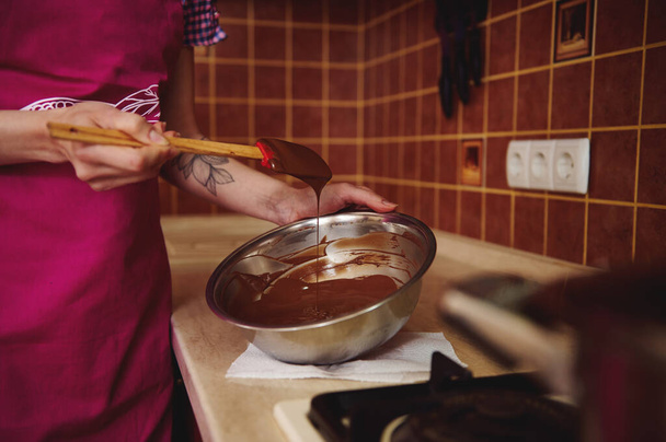 Крупный план неузнаваемой кондитерской в розовом фартуке, смешивающей расплавленную шоколадную массу в миске с деревянной лопаткой, стоящей у нее дома на кухне - Фото, изображение