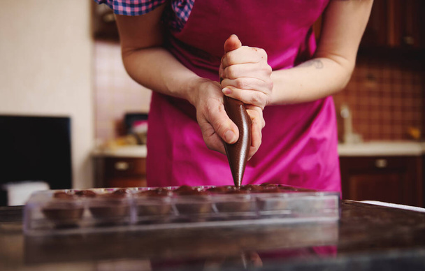 Vista cortada de chocolatier em avental rosa derramando enchimento de chocolate no molde de chocolate preparando luxuosos pralinos artesanais - Foto, Imagem