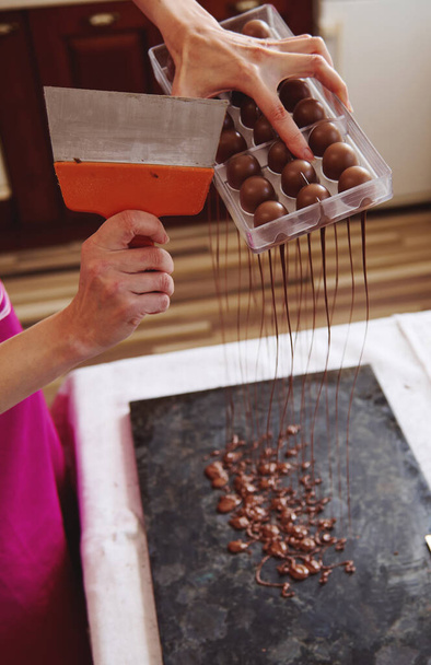Closeup van het gebruik van taart scraper om overtollige chocolade te verwijderen uit mallen in een marmeren tafel om snoep schelpen te maken. Het maken van luxe pralines en truffels thuis - Foto, afbeelding