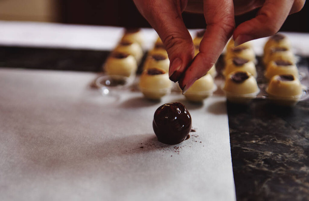 Focus sui tartufi di cioccolato su carta forno pergamena su uno sfondo di stampi di cioccolato e una mano di pasticcere cospargere cacao in polvere sulla caramella preparata - Foto, immagini