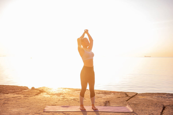 Young blonde woman in sportswear performs yoga asanas, exercises on the seashore at sunrise. The girl goes in for sports, works out breathing, meditates, performs Poses. Healthy mind in a fit body - Zdjęcie, obraz