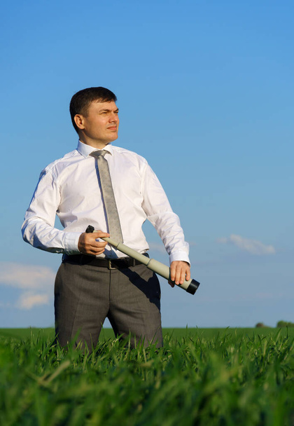 zakenman poseert met een verrekijker, hij kijkt in de verte en zoekt iets, groen gras en blauwe lucht als achtergrond - Foto, afbeelding