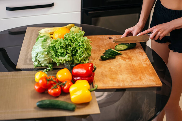 Pretty blond woman standing kitchen hold hand cut chopping cucumber products fruits vegetables wooden cutting board. Lady preparing breakfast lunch after training, want cook healthy food, copyspace. - Photo, Image