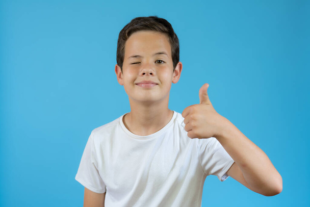 piękny młody chłopiec pokazując kciuk w górę gest na niebieskim tle - Zdjęcie, obraz