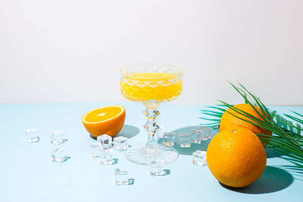 sklo s pomerančovou šťávou, pomeranče, kostky ledu, palmový list na modrém pozadí. - Fotografie, Obrázek