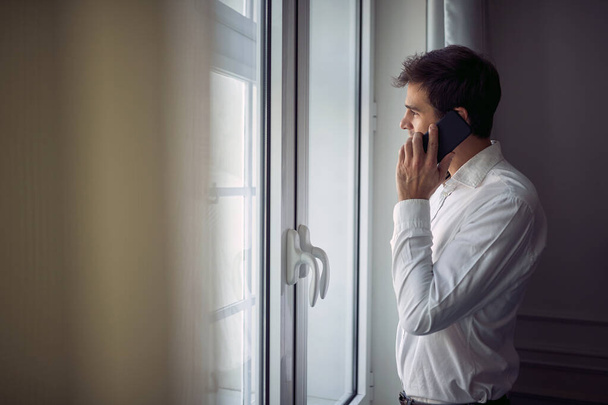 Όμορφος νεαρός λευκός άνδρας στέκεται με λευκό πουκάμισο, κοιτάζοντας έξω από το παράθυρο, μιλώντας στο κινητό του - Φωτογραφία, εικόνα