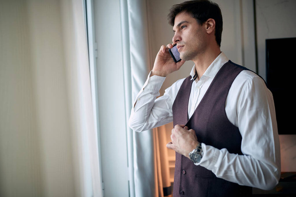 thoughtful young caucasian male talking on a cell phone by the window, fastening buttons on his vest with another hand - Fotoğraf, Görsel