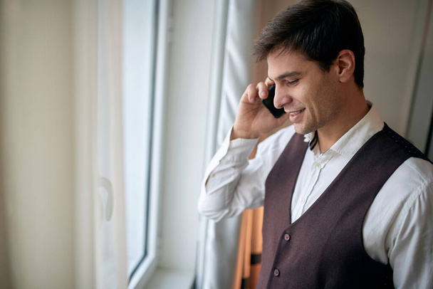 young caucasian male talking on a cell phone by the window, smiling, looking down, - Foto, Imagen