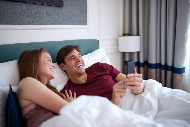 young caucasian adult couple laying in bed watching content on cell phone, laughing, having a good time - Фото, изображение