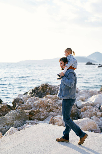 Smiling dad carries a little girl on his shoulders while walking along the pebble beach to the sea against the backdrop of green mountains and large boulders. Side view - Фото, зображення